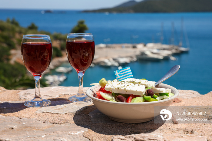 Two glasses of red wine and bowl of greek salad with greek flag on by the sea view, summer greek holidays concept.