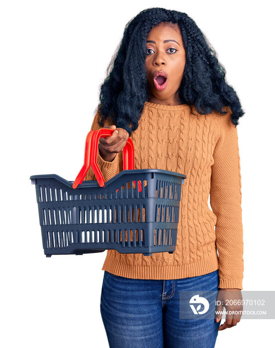 Beautiful african american woman holding supermarket shopping basket scared and amazed with open mouth for surprise, disbelief face