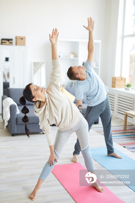 Young active man and woman standing on the floor with their left arms raised during home workout