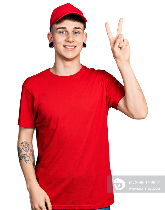 Young caucasian boy with ears dilation wearing delivery uniform and cap smiling with happy face winking at the camera doing victory sign. number two.