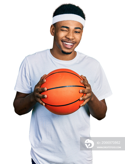 Young african american man holding basketball ball winking looking at the camera with sexy expression, cheerful and happy face.