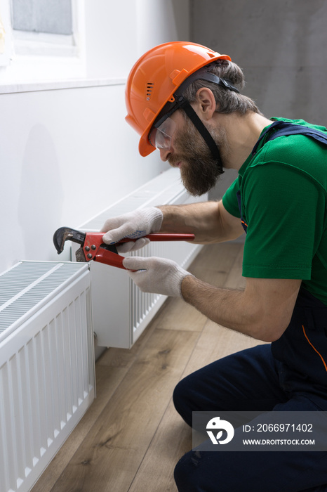 A male plumber installs a radiator in the heating system of an apartment. Guy in overalls and a gas wrench.