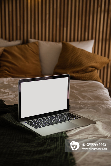 Laptop computer with blank screen in bed with pillows and blanket. Aesthetic minimal home, bedroom interior design template with mockup copy space. Work at home, quarantine self-isolation concept.