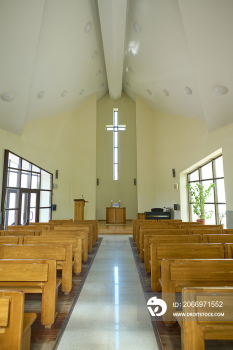 Interior of modern Catholic church with two rows of wooden benches for parishioners and long aisle leading to pulpit with cross above