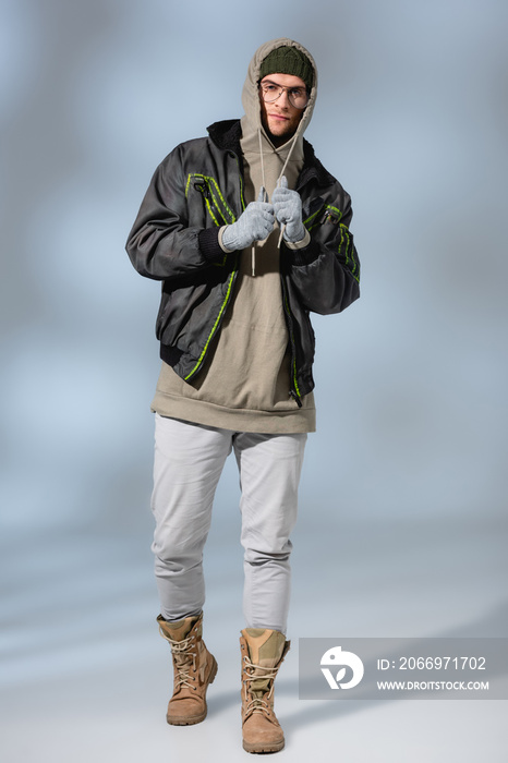 full length of young stylish man in hat, hoodie and anorak standing on grey