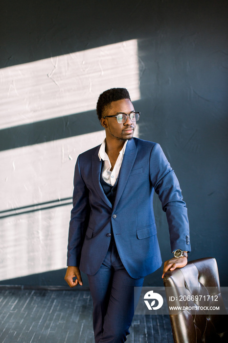 Young beautiful African American guy in a stylish suit standing in the office laying his hand on a chair