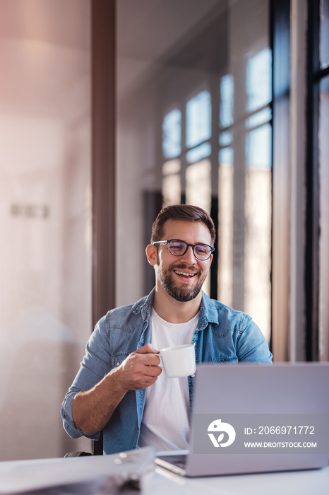 Handsome smiling young man sitting by window in office, drinking coffee while working on laptop.