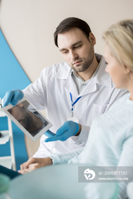 Bearded man in dentist office showing tablet to young client having consultation in cabinet.