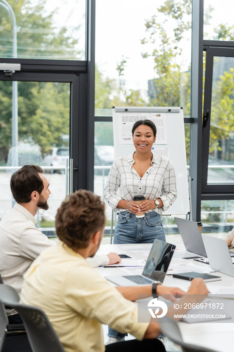 smiling multiracial woman with coffee to go talking to colleagues sitting at conference table