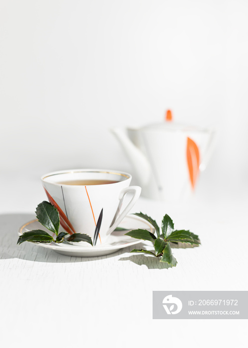 A branch and leaves of Yaupon holly lie near a porcelain cup and teapot on a table. Tonic herbal tea.