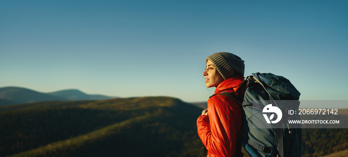 Side portrait woman hiker with backpack standing in sunset light among mountains and hills, panoramic view