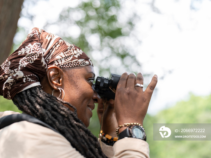 Smiling female hiker looking through binoculars in forest