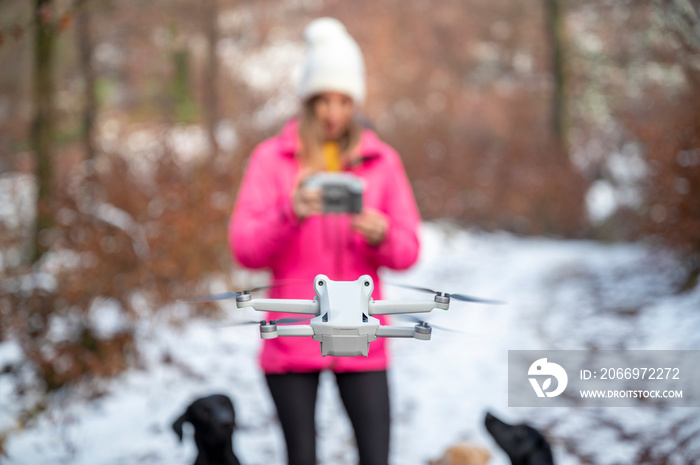 Young woman in pink winter jacket standing in snowy forest using a controller to direct and manage a drone