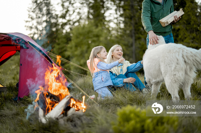 Young couple with a little girl have fun at campsite, traveling with tent on nature. Family spend summer time hiking in the mountains. Man carry firewoods