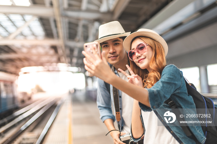 Happy Asian couple traveler holding a mobile phone in station and waiting for train in vacation time.Two Asian Tourists With Backpacks Train travel in Sightseeing City Thailand.