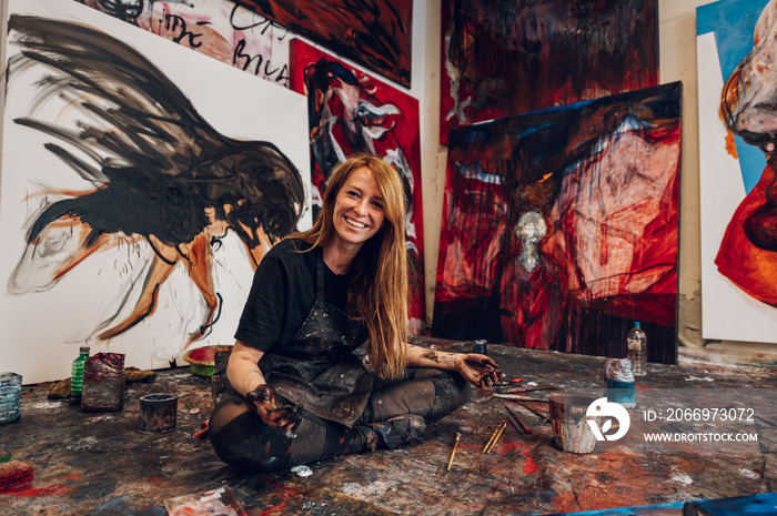 Young redhead woman painter sitting by her art masterpiece on a big canvas