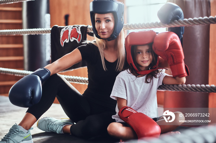 Young cute girl in helmet and her beautiful female boxing trainer are posing for photographer on the ring.