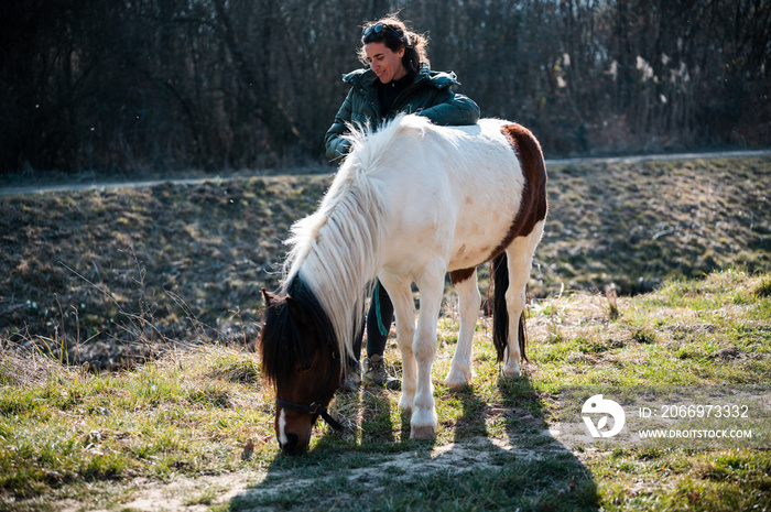 Beautiful no make-up young woman smiling and leaning on  her pony while it grazing,  enjoying sunny spring day outdoors.