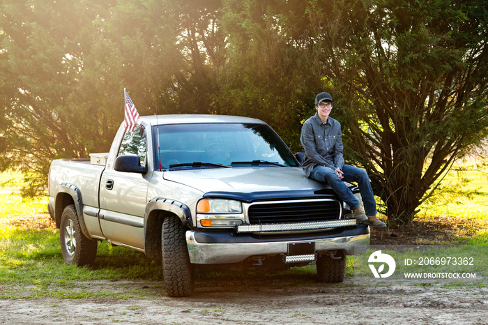 happy teenage boy from the south sitting on the front of his pickup truck outside in a rural area.