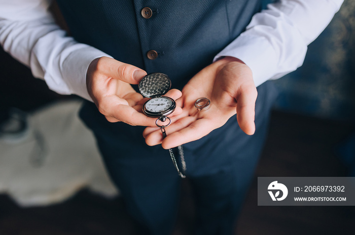 A man, a businessman in a suit holds in his hands a vintage, antique pocket watch and wedding rings close-up. The morning of the groom. Photography, concept, wedding. Clockwork.