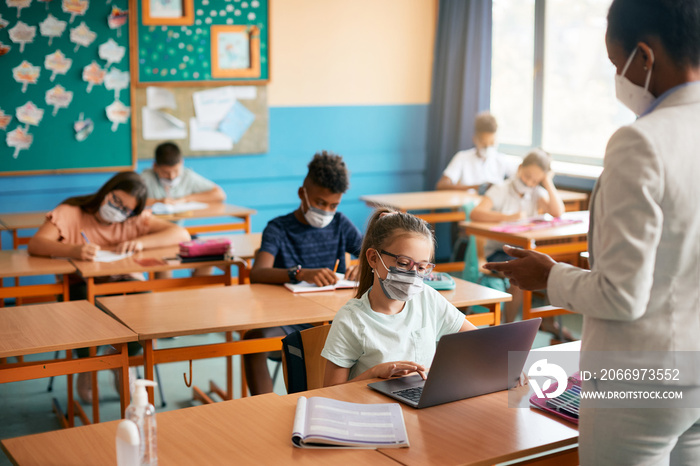 Schoolgirl wears face mask while e-learning on laptop during computer class in classroom.