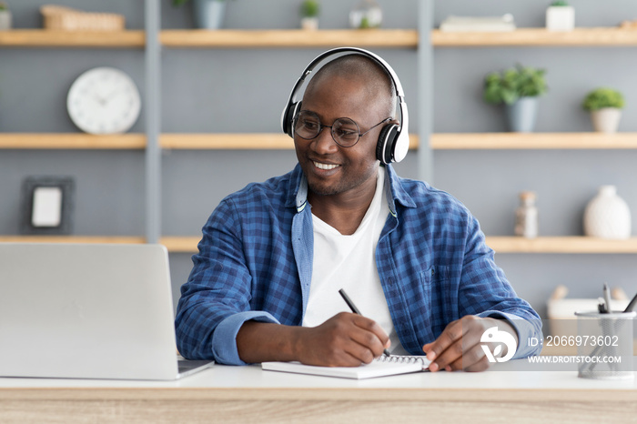 E-education. Happy black mature man using laptop and taking notes, listening online course