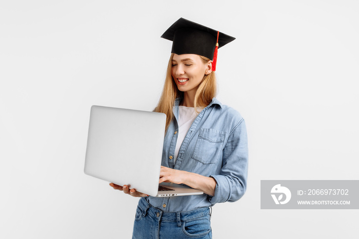 Happy excited beautiful graduate girl in a graduation cap on her head using a laptop while standing on a white background. Graduation, distance learning