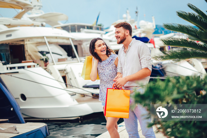 Happy young couple with shopping bags walking by the harbor of a touristic sea resort with boats on background