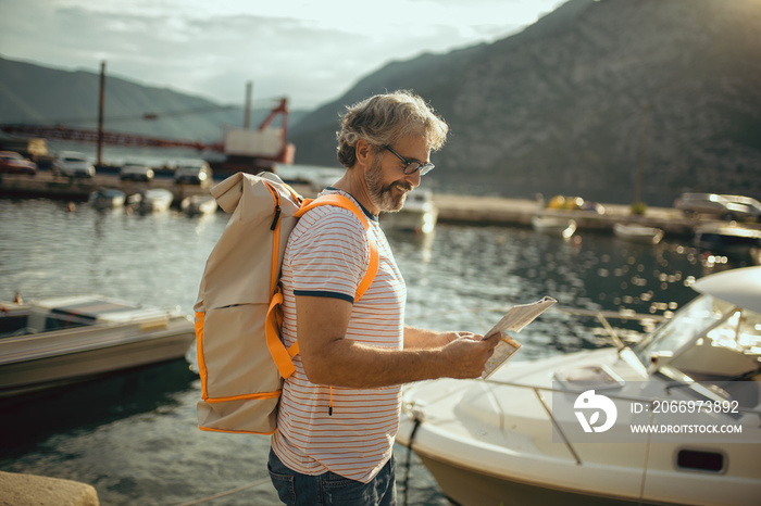 Smiling tourist mature man standing with map and backpack near the sea.