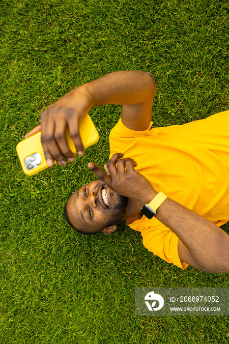 Happy young adult man laying on the grass, using smartphone, browsing internet. Joyful african american male relaxing, taking a break after workout outdoors, having a video call from friends, smiling