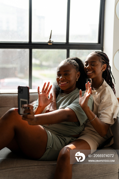 Lesbian couple waving while taking selfie on sofa at home