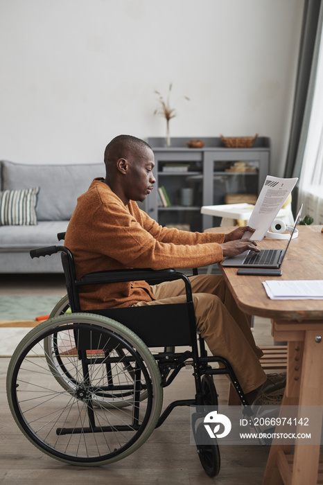 Vertical side view portrait of African-American man using wheelchair working from home in minimal grey interior, copy space