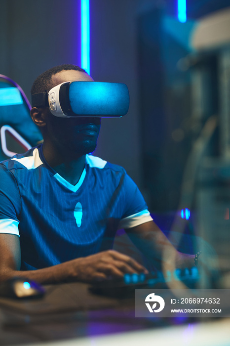 Serious concentrated black gamer in VR headset sitting at table and using computer while playing football video game