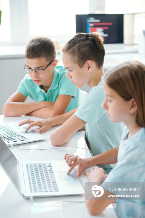 Row of serious cute schoolkids in casualwear sitting by desk in front of laptops