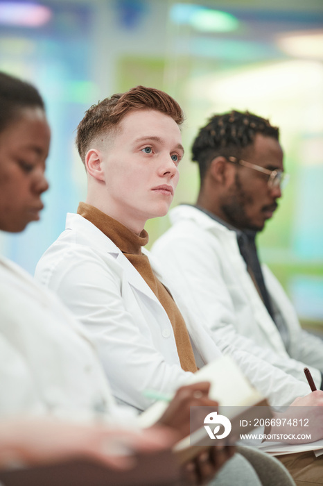 Vertical portrait of young man wearing lab coat while sitting in row in audience and listening to lecture on medicine in college