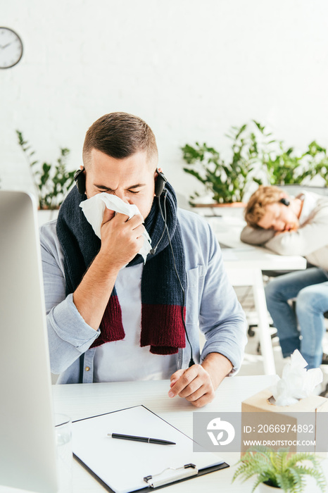 selective focus of sick broker sneezing in tissue near coworker in office