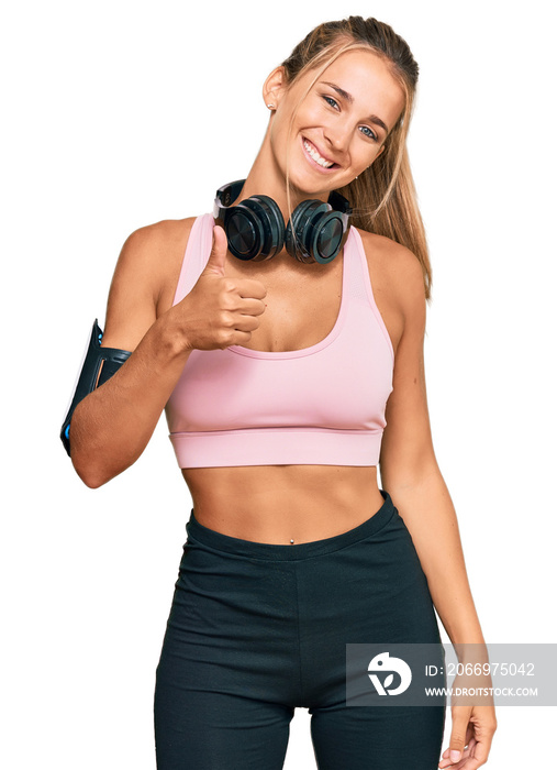 Young blonde woman wearing gym clothes and using headphones doing happy thumbs up gesture with hand. approving expression looking at the camera showing success.