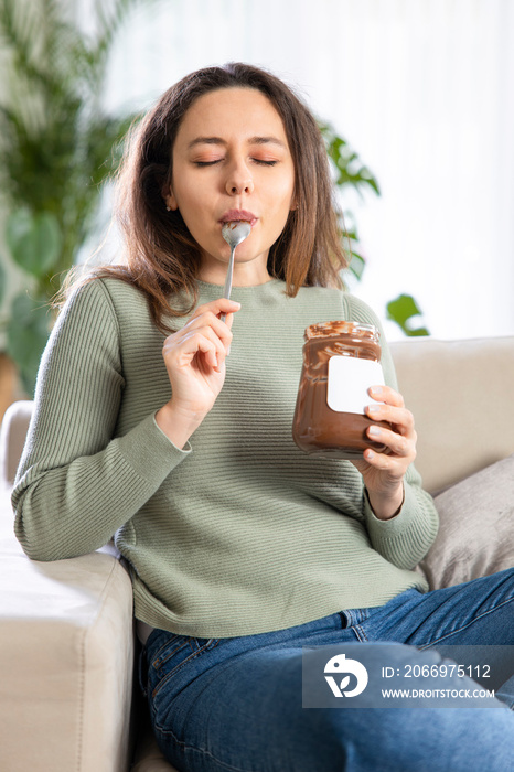 Young woman eating chocolate from a jar while sitting on on a couch.