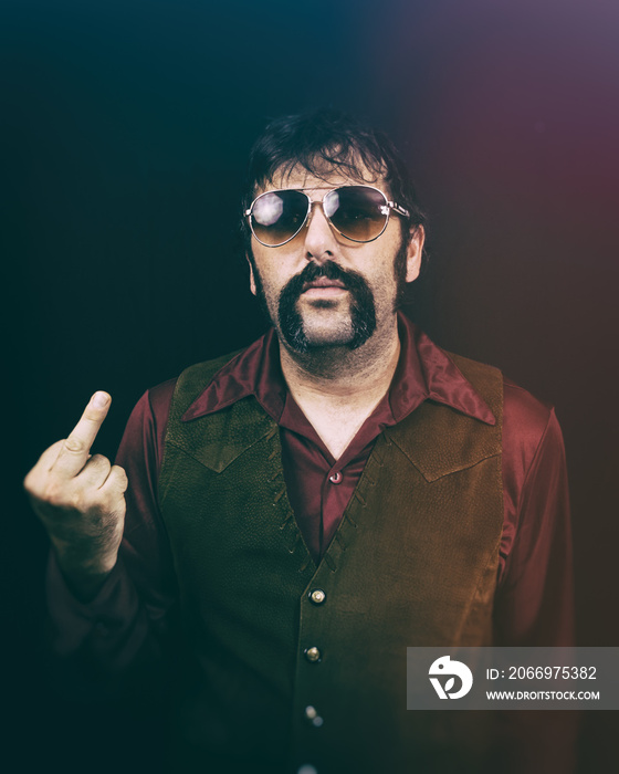 A man in authentic vintage 1970’s era clothing, including a vest, big collared shirt, mustache and sunglasses, making an obscene hand gesture.