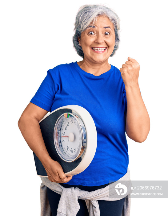 Senior woman with gray hair holding weight machine to balance weight loss screaming proud, celebrating victory and success very excited with raised arms