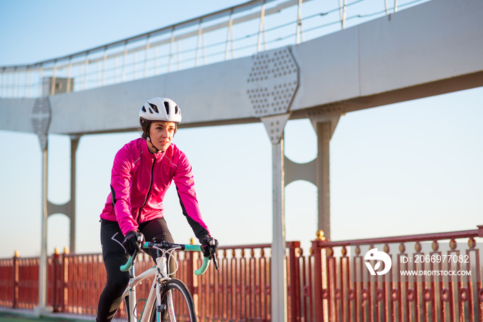 Young Woman in Pink Jacket Riding Road Bicycle on the Bridge Bike Line in the Cold Sunny Autumn Day. Healthy Lifestyle.