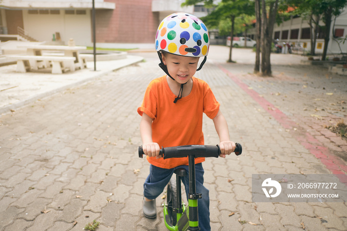 Close up of cute little Asian 3 years old toddler boy child wearing safety helmet learning to ride first balance bike on cement floor in sunny summer day, kid cycling at park