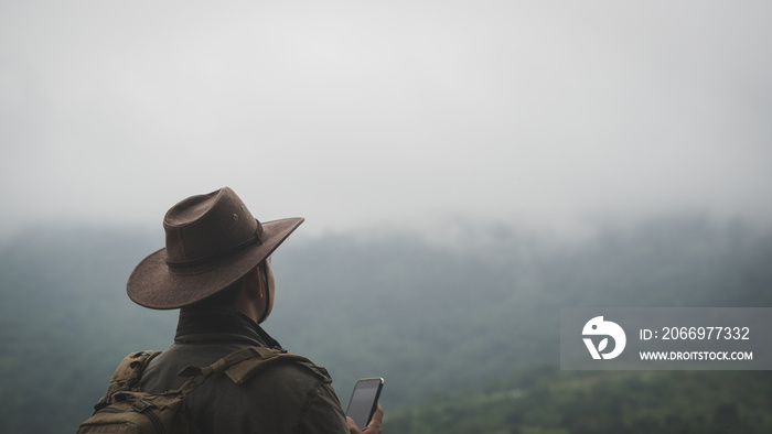 Freedom traveler man in hat carrying a backpack stands at the top of a mountain and using a smartphone on a foggy day.Adventure travel and success concept