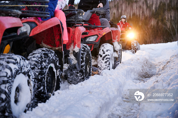 Close up of people riding red all-terrain vehicles down snowy hill. Quad riders driving quad bikes with black snowy wheels on snow-covered trail. Concept of winter activities and quad biking.