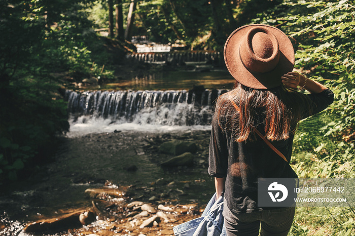 Stylish hipster girl traveling in woods, back view. Young woman in hat exploring in summer forest, looking at river. Travel and wanderlust concept. Space for text