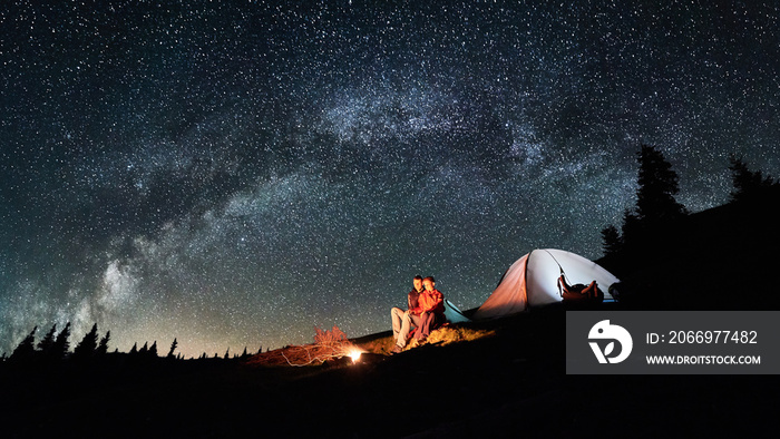 Night camping. Romantic couple tourists have a rest at a campfire near illuminated tent under amazing night sky full of stars and milky way. Astrophotography. Picture aspect ratio 16:9