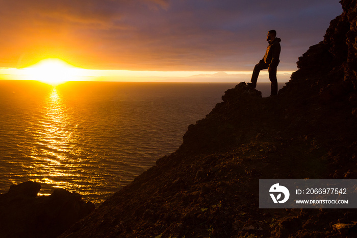 Lonely man standing on cliff edge rock staring at sunset with Teide mountain on background. Fearless person on mountain top enjoying spectacular twilight by the sea in Gran Canaria island, Spain