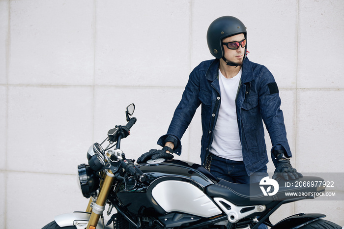 Serious motorcyclist in glasses and helmet leaning on vehicle and looking away