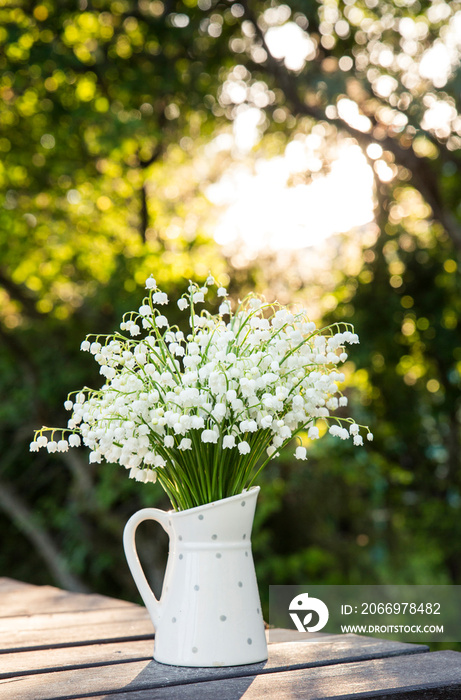 Bouquet of white flowers Lily of the valley (Convallaria majalis) also called: May bells, Our Lady’s tears and Mary’s tears in a white dotted jug shaped vase, outdoors on a table, trees on background.