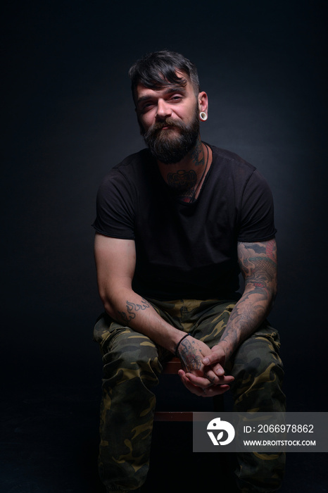 Brutal attractive bearded man with tattooed hands poses in black blank t-shirt and army pants seated on a chair